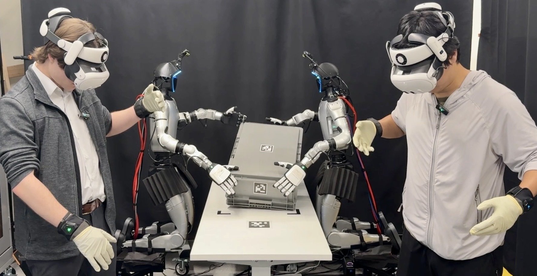 Two operators in Quest 3 headsets and gloves teleoperating two kneeling Unitree G1 humanoids holding a grey crate on a table